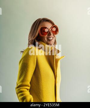 Trasudando fiducia e stile, una donna degli anni '40 che indossa un vivace cappotto giallo e occhiali da sole arancioni oversize sorride dolcemente, incarnando un senso di gioia Foto Stock