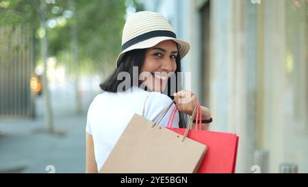 Primo piano di una donna sorridente con i capelli lunghi che indossa un cappello e tiene le borse della spesa sulla spalla. Lei guarda indietro con un'espressione gioiosa mentre Foto Stock