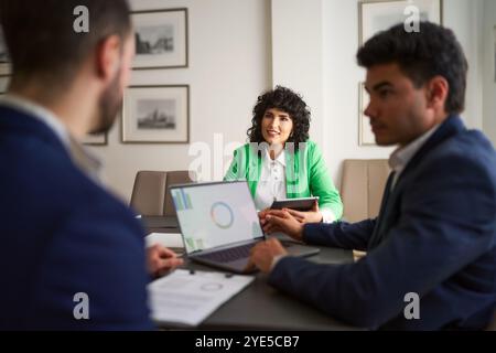 Tre persone sono sedute a un tavolo con computer portatili e documenti. Una donna è seduta al centro del tavolo e guarda il notebook. Gli altri due Foto Stock