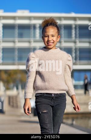 Una ragazza che indossa un maglione e un jeans passa davanti a un edificio. Ha un sorriso in faccia ed è felice Foto Stock