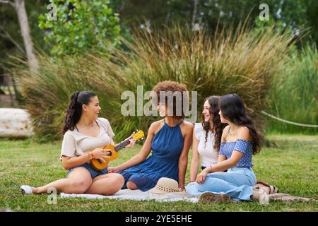 Quattro donne sono sedute su una coperta in un parco, una di loro gioca un ukulele. La scena è rilassata e amichevole, mentre le donne si divertono a vicenda Foto Stock