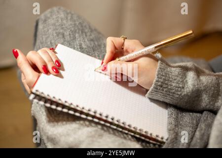 Una donna con unghie ben curate sta scrivendo intensamente in un taccuino vuoto utilizzando una penna elegante. Ama un momento di creatività nel suo accogliente spazio domestico. Foto Stock