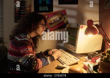 Persona che indossa un maglione colorato che scrive su un computer d'epoca in una stanza accogliente e poco illuminata, decorata con vari poster retrò sulla parete. Di fronte a una lampada calda, qualcuno immerso in una configurazione nostalgica Foto Stock