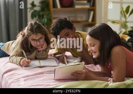 Tre bambini di diverse origini studiano e sorridono insieme mentre si sdraiano a letto con libri e quaderni che li circondano, creando un'atmosfera coinvolgente e vivace Foto Stock
