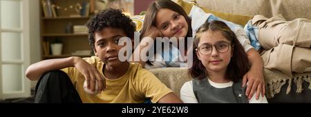 Tre bambini sdraiati sul divano, sorridenti e rilassanti durante il tempo libero al coperto. Mobili e scaffali visibili in un accogliente soggiorno Foto Stock