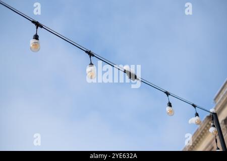 La neve poggia su luci decorative a corda appese sopra uno spazio all'aperto, adagiate su un cielo blu brillante, creando un'atmosfera invernale accogliente. Foto Stock