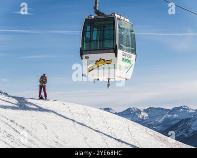 Una funivia si libra su una montagna innevata a Brianza, in Francia, mentre una persona con un abbigliamento colorato si erge sotto, osservando i paesaggi panoramici delle montagne Foto Stock