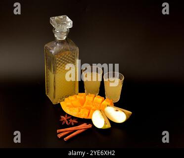 Liquore di mela-mango con spezie in vetro cristallo su sfondo nero, pezzi di frutta matura, cannella e anice. Primo piano. Foto Stock