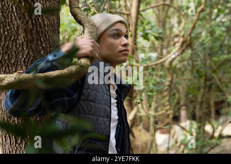 Giovane che esplora la foresta, tenendo il ramo dell'albero, indossando berretto e giacca Foto Stock
