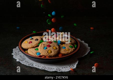 Gocce di cioccolato che cadono su un piatto nero con biscotti alla vaniglia rossi, blu, verdi e gialli su un tavolo nero Foto Stock