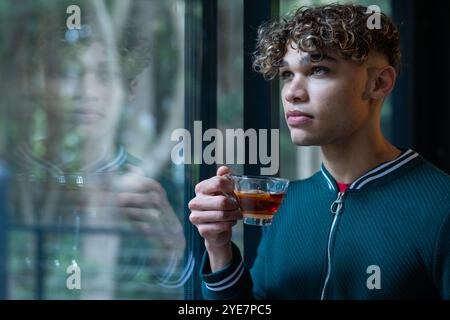 Giovane uomo riflessivo che gusta un tè alla finestra, contemplando momenti di pace, copia spazio, a casa Foto Stock