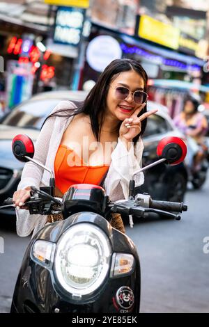 Una giovane signora tailandese reagisce alla telecamera con un grande sorriso mentre si fa strada lungo Soi Buakhao Pattaya, in Thailandia, con il suo scooter. Foto Stock