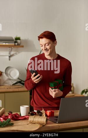 Un giovane allegro con un dono sorride mentre controlla il telefono in una cucina accogliente. Foto Stock