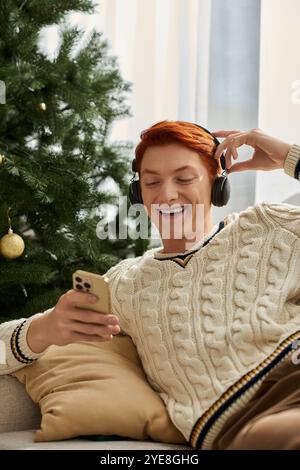 Un giovane si siede comodamente accanto a un albero di Natale, sorridendo mentre ascolta musica. Foto Stock