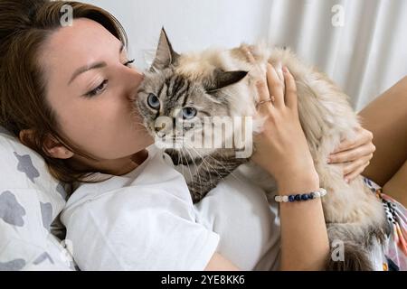 Una donna sta sdraiata su un letto con un gatto in grembo. Il gatto è soffice e ha gli occhi blu. La donna sta baciando il gatto in maschera neva, mostrando affetto Foto Stock