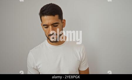 Il giovane ispanico con la barba ha un aspetto premuroso su uno sfondo bianco isolato, trasudando calma e fiducia in una semplice camicia bianca. Foto Stock