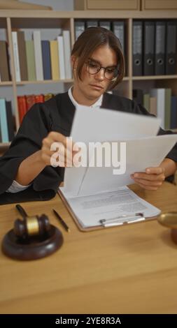 Giudice donna che legge documenti legali in un ufficio con un martelletto e libri sullo sfondo Foto Stock