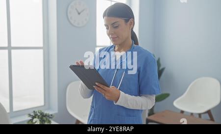 Una donna professionale infermiera in scrub controlla un tablet in una luminosa stanza d'ospedale, raffigurando la tecnologia sanitaria. Foto Stock