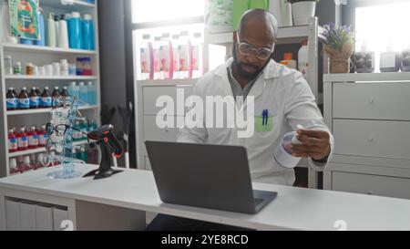 Bel giovane afroamericano con la barba che lavora in farmacia al coperto esaminando la bottiglia di medicina vicino al computer portatile e all'esposizione degli occhiali Foto Stock