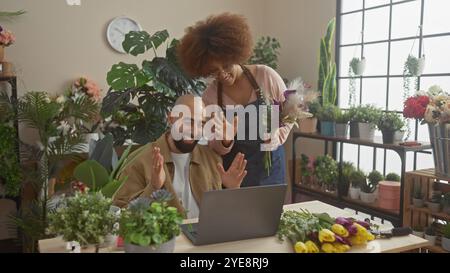 Una donna e un uomo, fioristi, ridono durante una videochiamata all'interno di un negozio di fiori circondato da piante. Foto Stock
