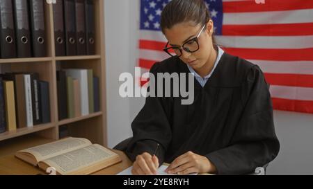 Giovane donna ispanica giudice che lavora alla scrivania in tribunale con bandiera americana in background, concentrandosi sui documenti legali. Foto Stock