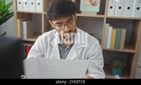 Giovane concentrato con barba in camice da laboratorio che legge un documento all'interno di una clinica. Foto Stock