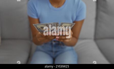 Donna che conta i soldi in casa su un divano, che mostra banconote da cento dollari mentre siede in un abito casual in un salotto Foto Stock