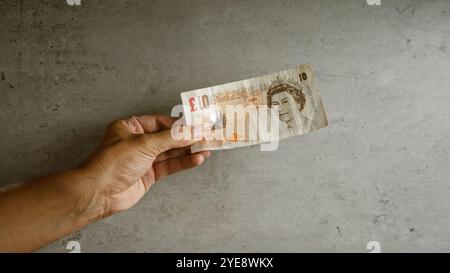 Mano di un uomo che tiene una banconota da dieci sterline su un fondo di cemento che mostra la valuta britannica Foto Stock