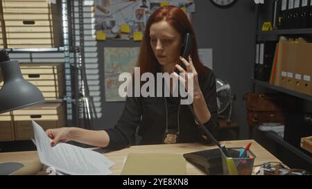 Detective rossa concentrata che esamina documenti e parla al telefono in un ufficio disordinato. Foto Stock