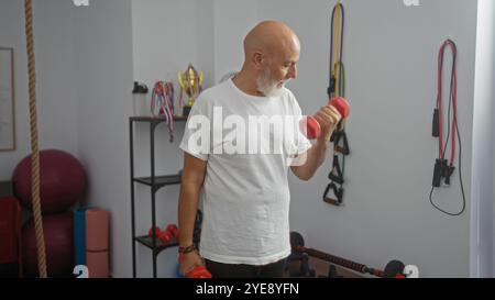 Uomo di mezza età con manubri che sollevano la barba in una palestra ben attrezzata con vari accessori per il fitness e medaglie sullo sfondo, trasportando un acti Foto Stock