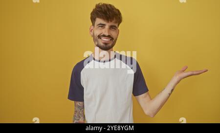 Un giovane sorridente con barba e tatuaggi estende la mano su uno sfondo giallo della parete in una posa informale. Foto Stock