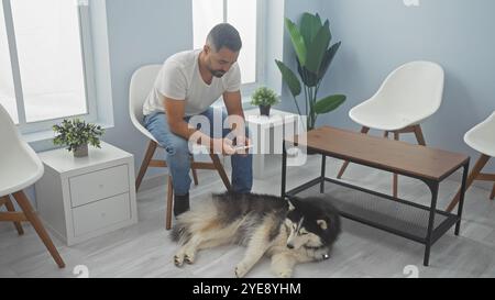 Bell'ispanico che controlla il telefono nella hall della clinica veterinaria con un cane husky rilassato. Foto Stock