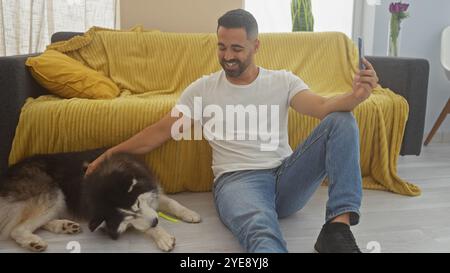 Un giovane allegro scatta un selfie con il suo cane da husky in un accogliente salotto, incarnando uno stile di vita domestico informale. Foto Stock