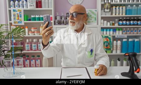 Uomo di mezza età con la barba che fa una videochiamata in farmacia mentre tiene in mano una bottiglia di farmaci, circondato da vari prodotti e occhiali in dispensa Foto Stock