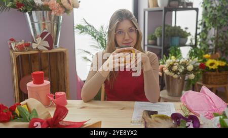 Una giovane donna bionda si gode una tazza in un intimo negozio di fiori circondato da mazzi e avvolgimenti creativi. Foto Stock