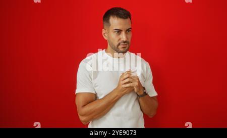 Bellissimo giovane ispanico con una barba in piedi su un vivace sfondo rosso con una camicia bianca che stringe le mani Foto Stock