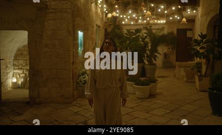 Una giovane donna ispanica cammina attraverso le strade splendidamente illuminate di polignano a mare, puglia, italia di notte, circondata da piante e affascinanti ancien Foto Stock