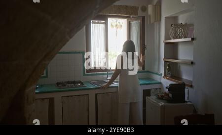 Una giovane donna ispanica in una vecchia cucina italiana si affaccia fuori dalla finestra con un arredamento rustico e un'atmosfera accogliente. Foto Stock