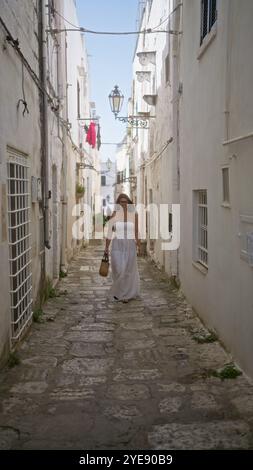 La giovane donna ispanica percorre una bellissima stradina nel centro storico di ostuni, puglia, italia, europa, tenendo in mano una borsa e indossando una dres bianca Foto Stock