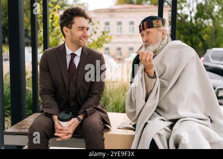 Uomo d'affari sorridente e empatico che ascolta il vecchio senzatetto seduto sulla panchina, che rappresenta il divario crescente tra ricchi e poveri nella società. Foto Stock
