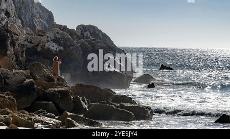 Woman si erge sulle rocce a fotografare l'oceano e sulla costa frastagliata delle spiagge di Sagres in Portogallo. Sagres si trova all'estremo occidentale... Foto Stock