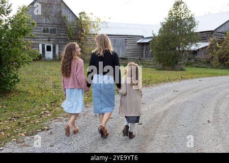 Vista da dietro di una madre che si tiene per mano con le sue due figlie mentre camminano lungo una strada di ghiaia verso un vecchio edificio rurale in legno... Foto Stock