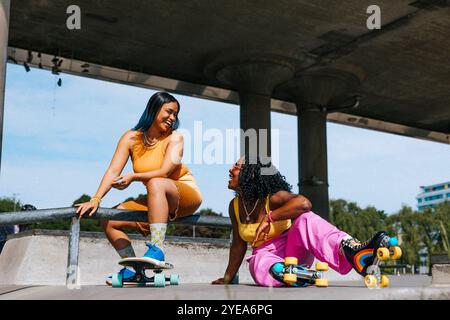 Amiche felici che indossano pattini a rotelle sedute vicino al cavalcavia nelle giornate di sole Foto Stock