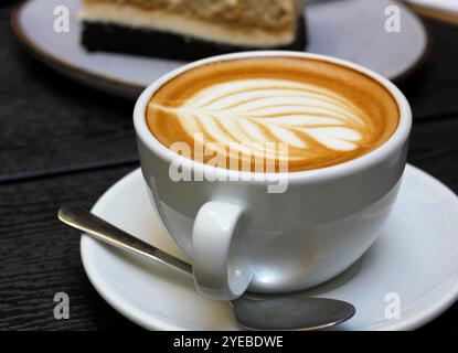 Tazza bianca di caffe latte con rosetta latte art, cucchiaino e piattino, torta tiramisù sullo sfondo, tavolo di legno scuro. Primo piano di caffè e torta Foto Stock