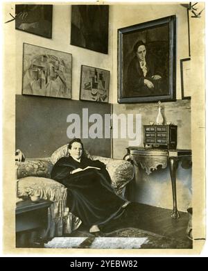 Gertrude Stein seduta su un divano nel suo studio di Parigi, con un suo ritratto di Pablo Picasso, e altri dipinti d'arte moderna appesi al muro dietro di lei. 1930. Foto Stock