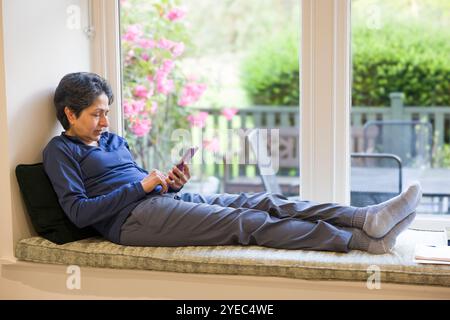 Donna asiatica indiana di mezza età che si rilassa seduta su un posto vicino alla finestra con i piedi in alto, guardando lo schermo del telefono con un giardino inglese sullo sfondo Foto Stock