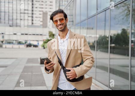 Felice bel giovane ragazzo del medio Oriente con occhiali da sole e casual che va in ufficio, cammina per strada, usa il telefono, controlla la posta elettronica, usa il cellulare aziendale A. Foto Stock