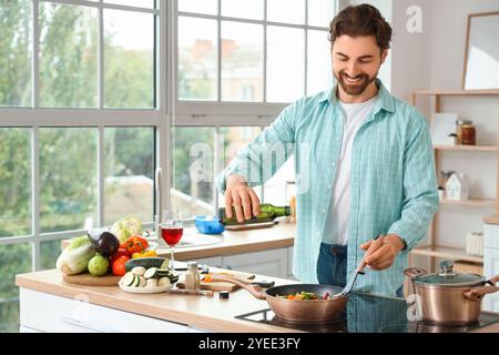 Bell'uomo barbuto che versa olio su verdure fritte in cucina Foto Stock
