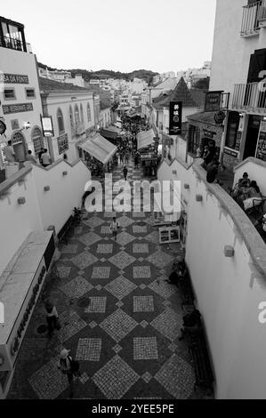 Una vivace strada nella città vecchia di Albufeira, catturata in bianco e nero, che mostra l'atmosfera vivace e la passerella piastrellata Foto Stock