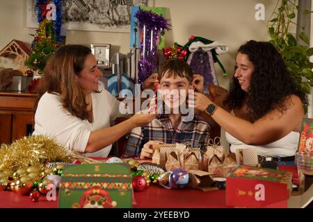 Una scena natalizia con due donne che decorano un giovane con ornamenti natalizi. Il tavolo è decorato con decorazioni colorate, scatole regalo e h Foto Stock